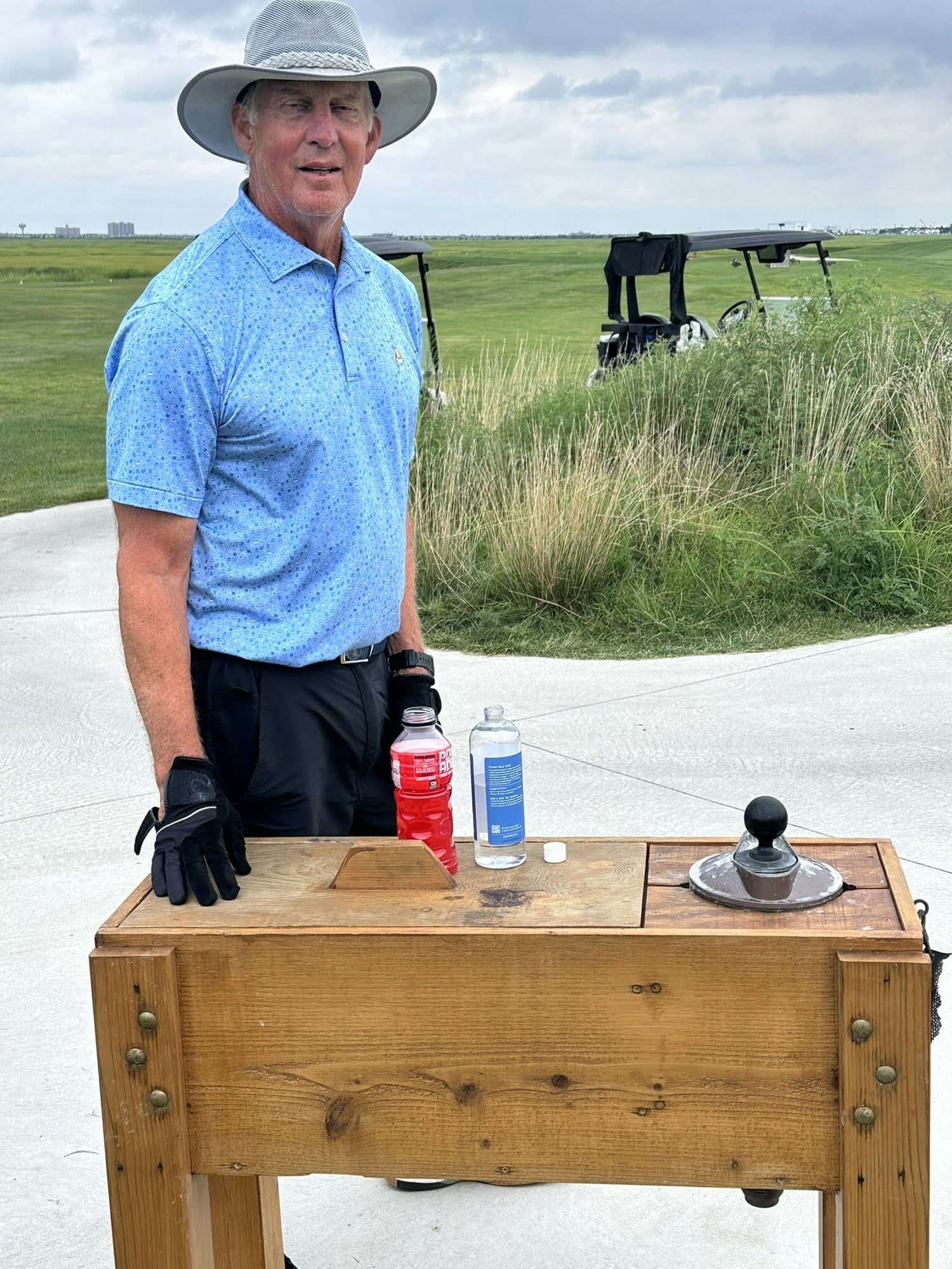A golfer in a sun hat at a tee box, drinks lined up on the ball washer